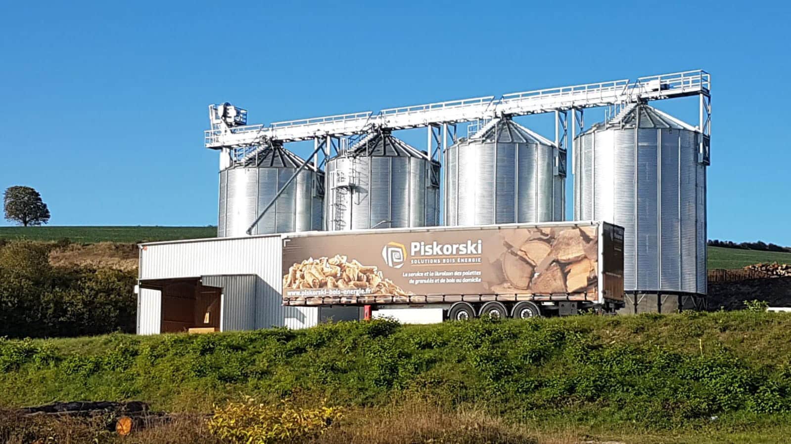 un camion pellet garé devant quatre silos a fond conique avec passerelle supérieure