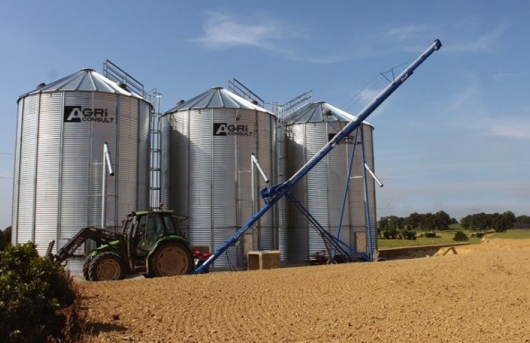 un tracteur avec vis sur chariot devant 3 silos à grain de stockage