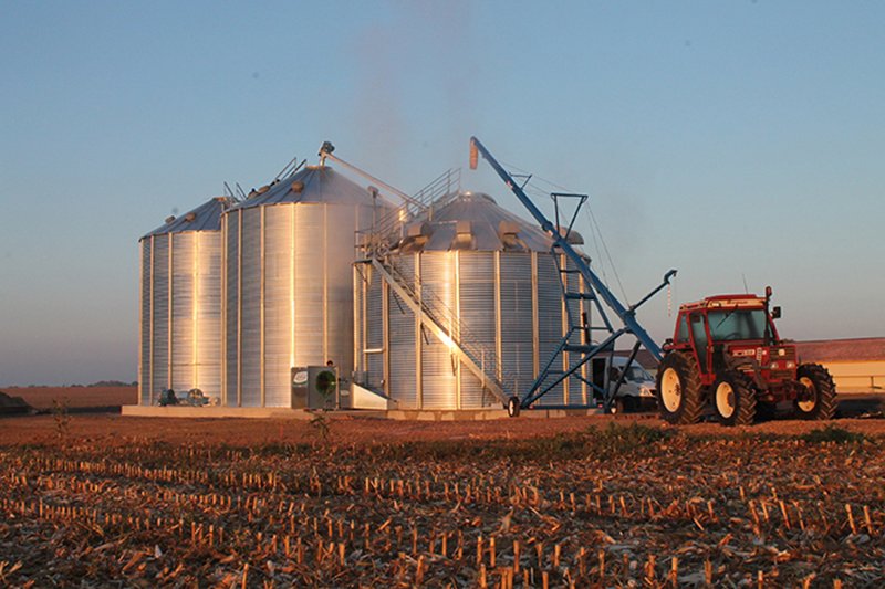 tracteur et vis à grain sur chariot chargeant une cellule sécheuse et deux silos de stockage au soleil couchant